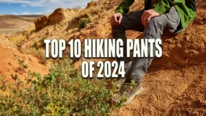 A hiker adjusting his gray pants while sitting on a sandy hill, with a backdrop of a rugged mountainous landscape under a blue sky.