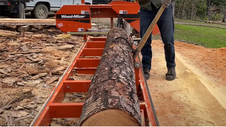 Wood-Mizer LT15 sawmill in action. The sawmill, painted in vibrant orange with a black stripe, is cutting through a large log. A person operates the sawmill using a long pole. The background features trees and a dirt road, emphasizing the practicality and functionality of this sawmill