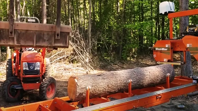 Wood-Mizer LT15 sawmill situated in a forest. The sawmill is orange and has a log on its bed. In the background, there’s a red tractor with a front bucket.