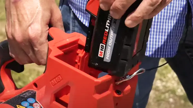 Person, wearing a blue and white checkered shirt, inserting a black “M18 REDLITHIUM HIGH OUTPUT” battery into a red Milwaukee M18 FUEL chainsaw