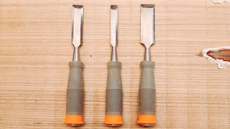 Three chisels with varying sizes on a wooden surface, with gray and orange handles.