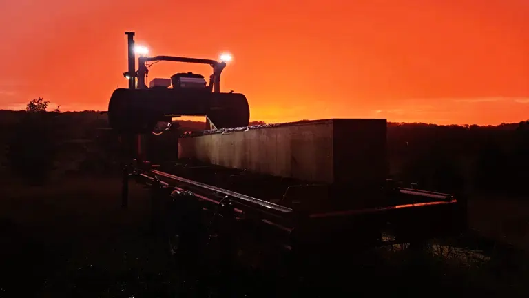 Norwood LumberPro HD36V2 sawmill against the backdrop of a red sunset. The sawmill is black, equipped with a large blade and a long bed. It stands prominently in the foreground, while the trees and the vibrant sunset form the background. Lights on the sawmill are illuminated, creating a striking contrast against the evening sky.