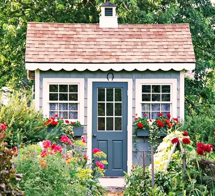 Charming garden shed with gray walls, white trim, and a cedar shingle roof, surrounded by colorful flowers.