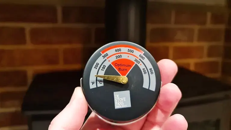 Hand holding a black and orange thermometer labeled ‘Optimum Burn’ and ‘UK Stove Fans’ in front of a brick woodstove.