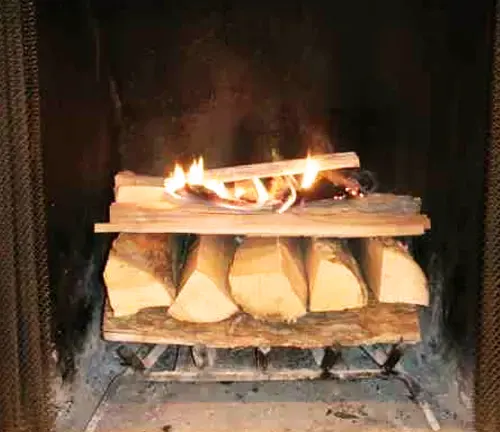 Fire burning on top of stacked large logs in a black cast iron wood stove, against a brick wall.
