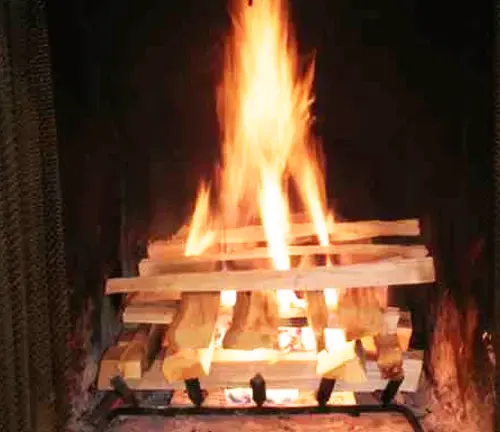 Fire burning on top of stacked small logs in a black cast iron wood stove, against a brick wall.