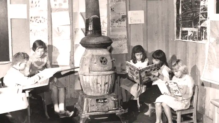 Black and white photo of a potbelly wood stove with children reading around it.