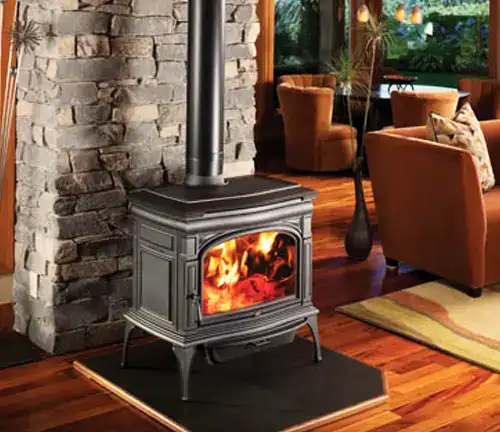 Lopi Cape Cod wood stove with fire, on stone hearth in a living room with orange armchairs and large windows.