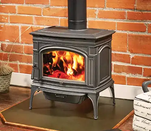 Black Lopi Cape Cod wood stove with vibrant fire, on dark hearth pad against red brick wall.