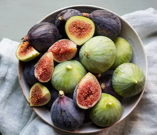 The image showcases a bowl of fresh figs in various colors and sizes. The figs are housed in a white bowl with a blue rim, which is placed on a white cloth with a blue stripe. The background is a light blue color, providing a soothing backdrop that complements the vibrant colors of the figs. The image beautifully captures the freshness and diversity of the figs, making it a delightful visual treat.