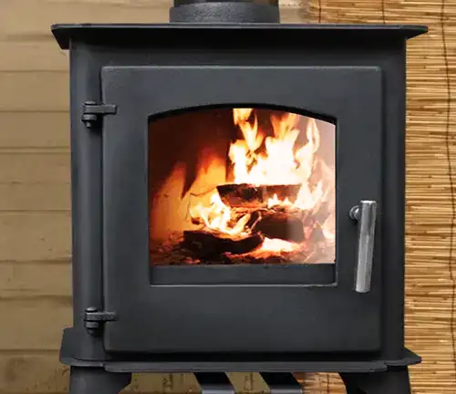 Capybara Mini Wood RV Stove with a fire burning inside, placed against a bamboo wall