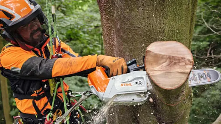 Tree surgeon in orange safety gear using a chainsaw to cut a tree trunk.