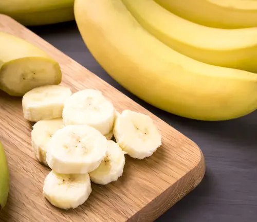 Bunch of ripe bananas and sliced bananas arranged in a circular pattern on a wooden cutting board