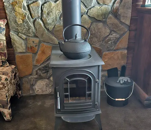 Wood burning stove with a kettle on top, set against a stone wall.