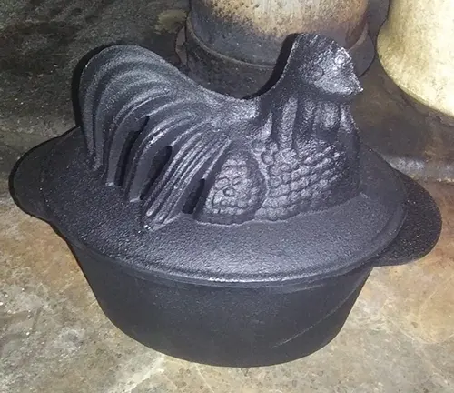 Cast iron pot with a rooster-shaped lid on a stone surface.