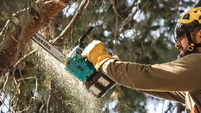 Person in yellow helmet using a chainsaw to cut a tree branch.