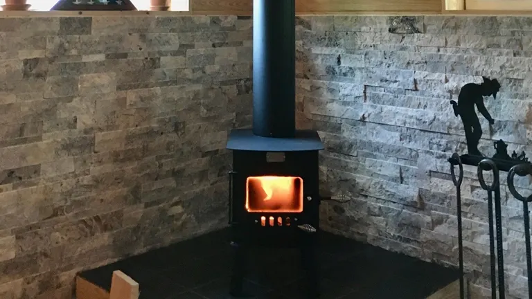 Black wood-burning stove with a burning fire, against a stone wall.