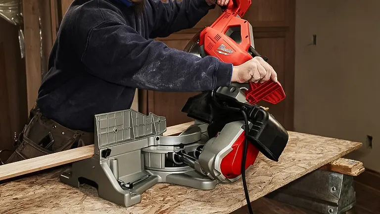 Person in blue sweatshirt using a red and gray Milwaukee 6955-20 12” Dual-Bevel Sliding Compound Miter Saw to cut a wooden plank on a workbench