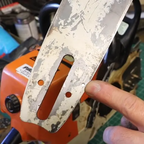 Close-up of a person’s finger pointing at a dirty or rusted chainsaw blade with an orange body, surrounded by various tools and equipment