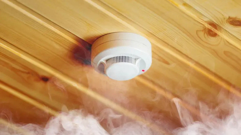 Smoke detector on a wooden ceiling with smoke around it.