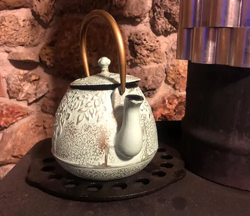 White ceramic teapot with a brass handle and spout on a black metal trivet against a brick wall.