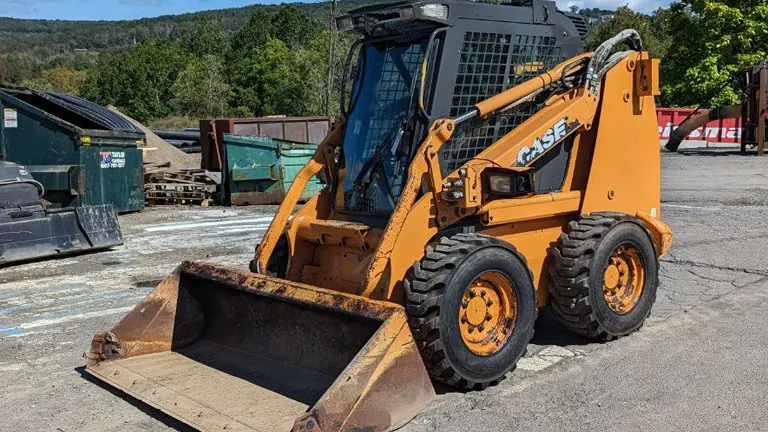 Orange CASE skid steer loader parked in a lot with dumpsters and trees in the background.