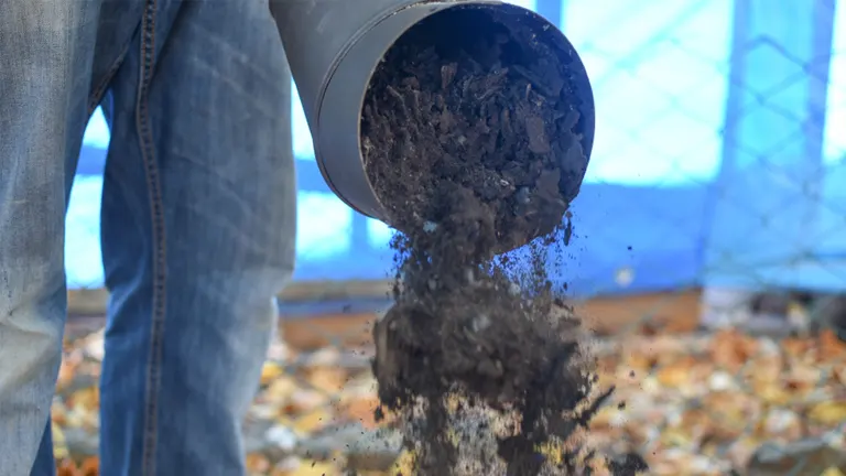 Person in blue jeans and gray shirt, emptying a rusted metal bucket of soil onto a pile of autumn leaves.