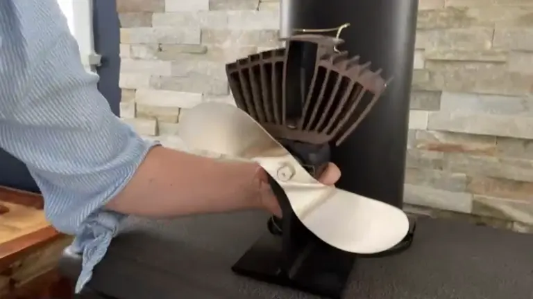 A person holding a Caframo Wood Stove Fan in front of a stove. The fan is cream-colored with black blades. In the background, there’s a modern wood-burning stove with a cylindrical shape and matte black finish. The wall behind the stove is adorned with rectangular stone tiles in various shades of grey