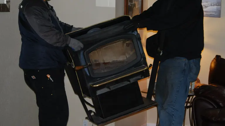 Two people in dark clothing moving a black wood-burning stove in a living room.