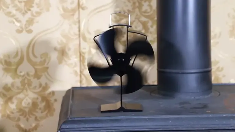 Tomersun Heat Powered Stove Fan on a wood stove. The fan has four black blades and is designed to distribute heat across the room. A metal flue pipe extends upwards from the wood stove, indicating it’s actively connected for venting smoke and gases. The wall behind the stove and fan has an ornate, patterned wallpaper. The base of the fan is metallic, ensuring it can withstand the heat from the stove below it