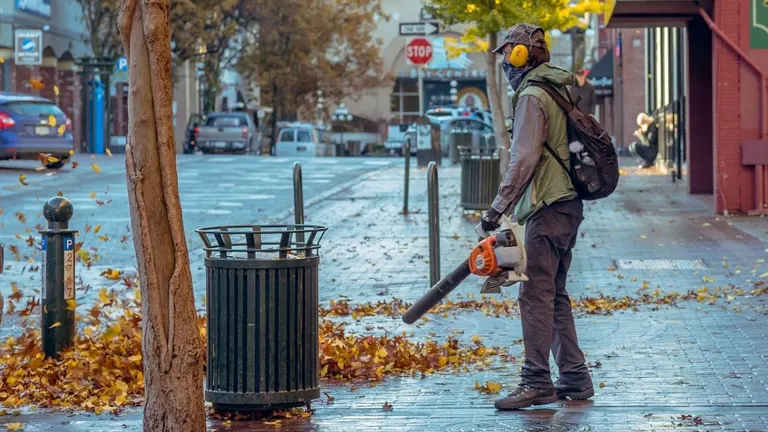 Person using a leaf blower on a sidewalk with fallen leaves