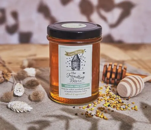 Jar of ‘The Travelling Bee Co.’ honey with honeycomb and bee pollen on a table.