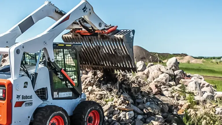 Bobcat S650 skid steer loader with a red grapple bucket moving rocks.
