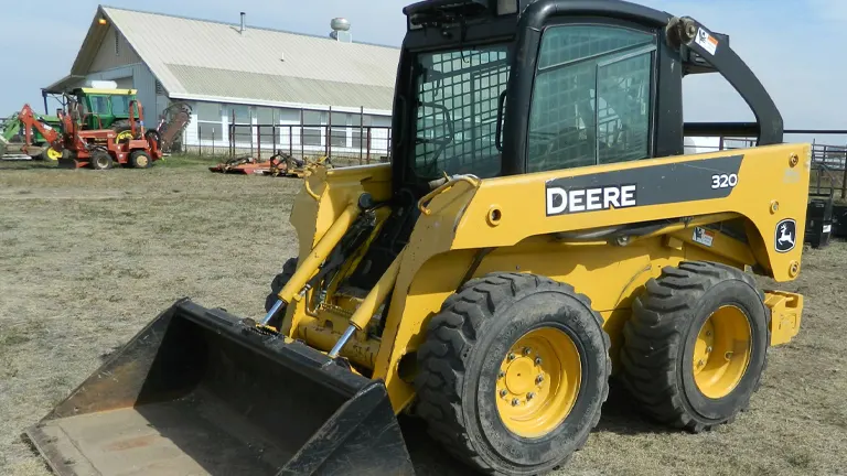 Yellow John Deere 320 skid steer loader with a fork attachment in a construction site.