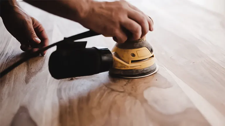 Person using a belt sander on a light brown hardwood floor