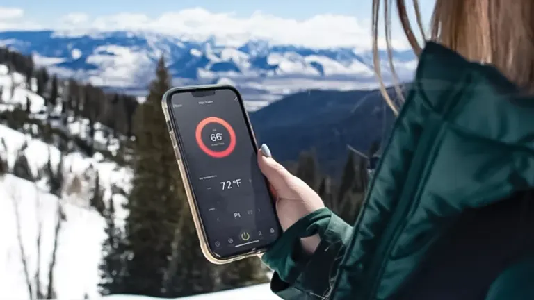 Person holding a phone displaying a temperature app in front of a snowy mountain landscape.