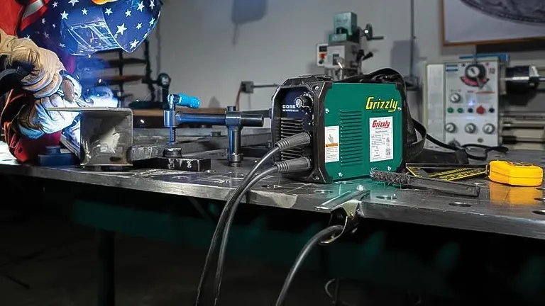 Grizzly G0880 85A Stick Welder on a workbench, with a blurred background of a welder at work.