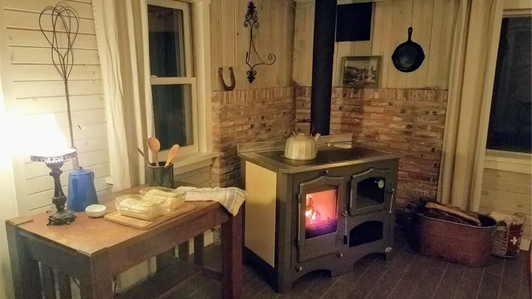 Cozy cabin interior with wood-burning stove, rustic decor, wooden table with blue lamp, and vase of flowers.