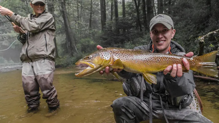 two individuals in outdoor clothing, one standing in the water with fishing equipment and the other kneeling in the water presenting a large, spotted trout, in a shallow river within Pisgah National Forest