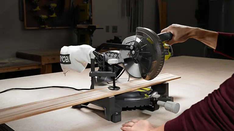 Person using Ryobi TS1144 9 Amp Corded 7-1/4” Compound Miter Saw to cut a wooden plank in a workshop