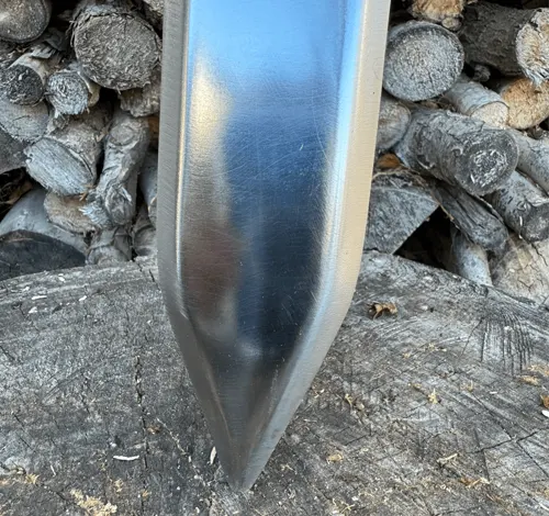 Polished sword with a 60-degree pointed tip resting on a wooden surface, with stacked logs in the background