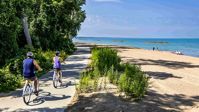 Two people biking on a trail at Presque Isle State Park with a sandy beach and blue lake in the background