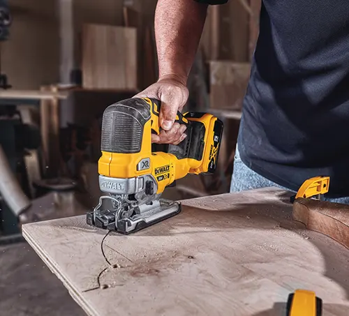A person using a yellow and black DeWalt jigsaw to cut through a wooden board in a workshop