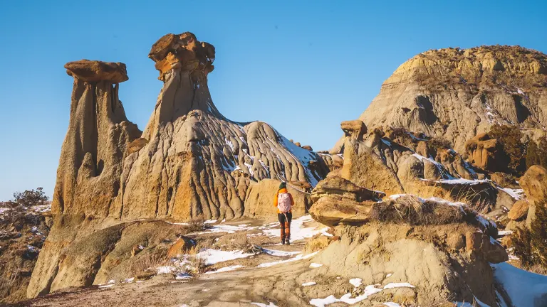 A person hiking amidst the unique rock formations of Makoshika State Park with snow patches on the ground