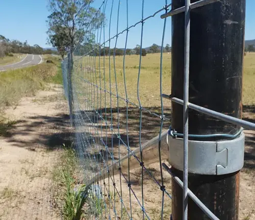 Close-up view of a metal fence post with attached wire fencing along a rural road