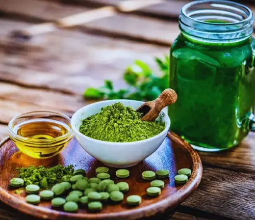 various Moringa products on a wooden tray. It includes green Moringa powder in a bowl, golden Moringa oil in a glass container, green Moringa tablets scattered on the tray, and a green Moringa-based drink in a mason jar.