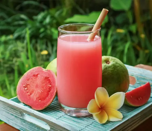 A glass of pink guava juice with a straw, surrounded by fresh guavas and a flower, on a wooden surface with greenery in the background