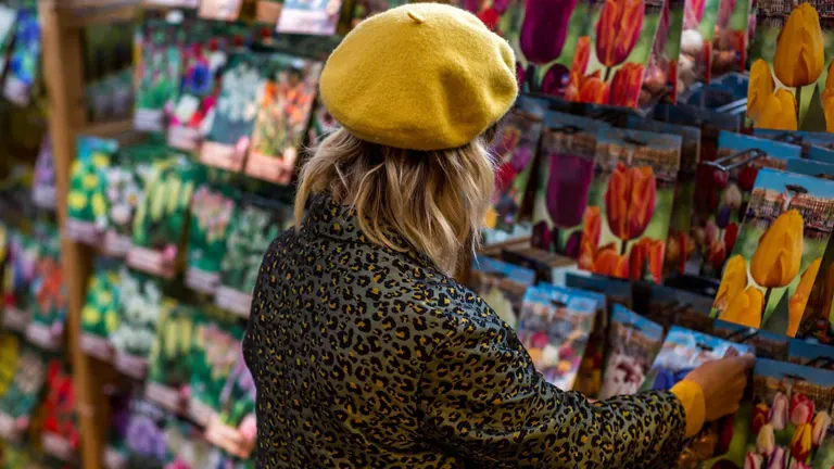 A person in a yellow beret and leopard print jacket browsing a wide variety of seed packets in a store, indicating the importance of seed selection for garden space
