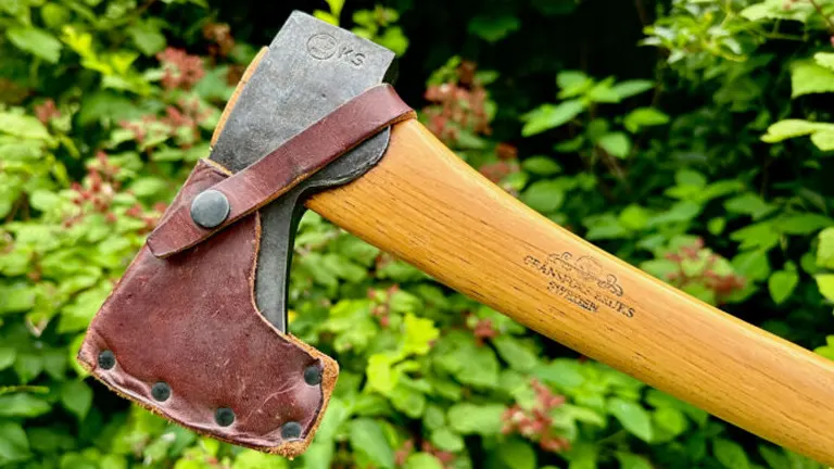 Camping hatchet with a wooden handle and metal head, partially covered by a worn leather sheath, against a green foliage backgroun