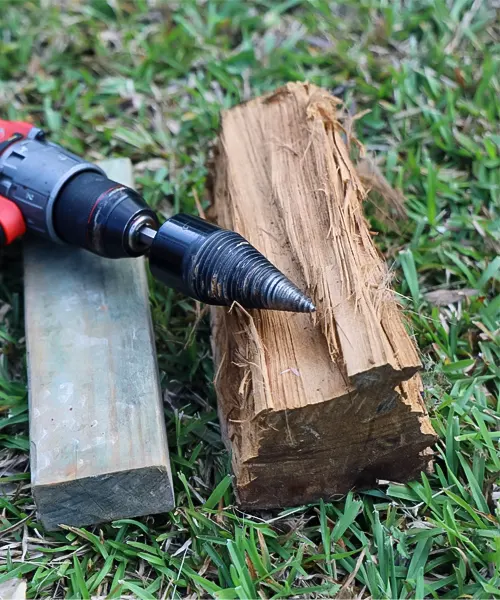 A drill with a firewood log screw splitter bit inserted into a piece of wood on a grassy ground